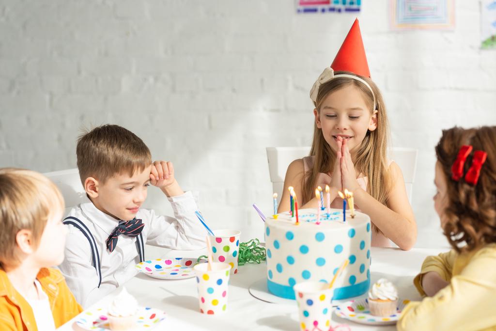 αξιολάτρευτο παιδί κάνει παρακαλώ χειρονομία και να κάνει την ευχή, ενώ κάθονται στο τραπέζι με τους φίλους κατά τη διάρκεια γιορτή πάρτι γενεθλίων - Φωτογραφία, εικόνα