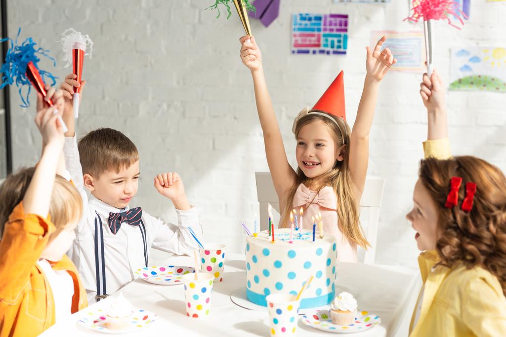 gelukkige kinderen zitten aan tafel met taart en juichen met partij hoorns tijdens de verjaardag viering - Foto, afbeelding