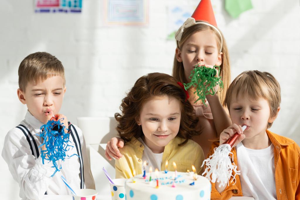 niños sentados en la mesa con pastel y animando con cuernos de fiesta durante la celebración del cumpleaños
 - Foto, Imagen