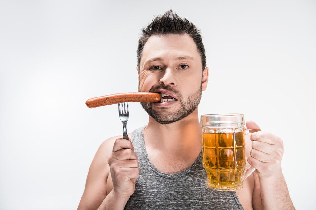 παχουλός άνθρωπος δάγκωμα λουκάνικο και κρατώντας ένα ποτήρι μπύρας απομονωθεί σε λευκό - Φωτογραφία, εικόνα