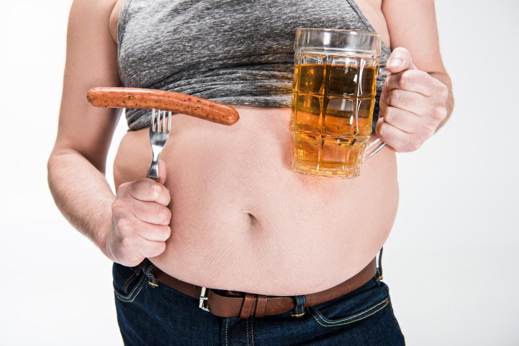 przycięte widok z nadwagą człowiek pokazując brzuch i trzymając szklankę piwa z grillowaną kiełbasą na białym tle - Zdjęcie, obraz