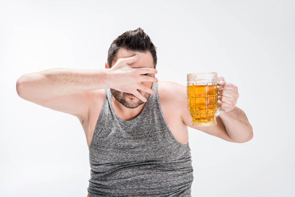 Übergewichtiger Mann bedeckt Gesicht mit Hand und hält Glas Bier auf weißem Grund - Foto, Bild