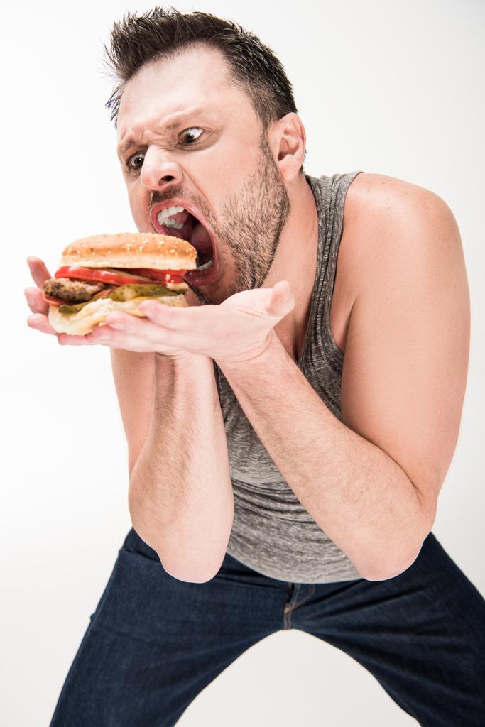 Übergewichtiger Mann macht Gesichtsausdruck, während er mit Burger auf Weiß posiert - Foto, Bild