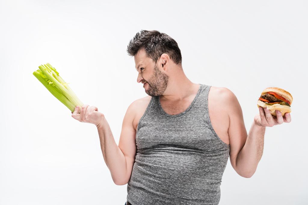 skeptical overweight man holding celery and burger isolated on white  - Photo, Image