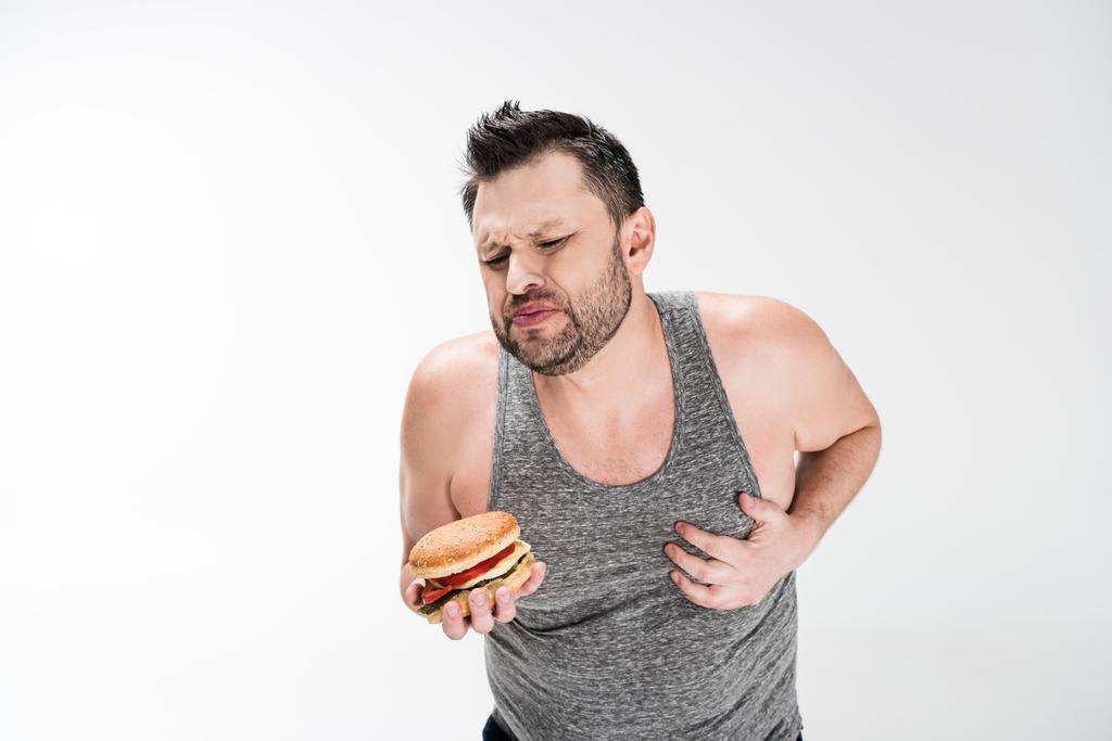 hombre con sobrepeso sosteniendo hamburguesa y tocando el pecho en blanco con espacio de copia
 - Foto, Imagen