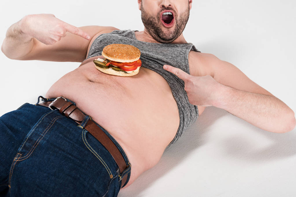 abgeschnittene Ansicht eines aufgeregten übergewichtigen Mannes, der mit den Fingern auf Burger am Bauch auf Weiß zeigt - Foto, Bild