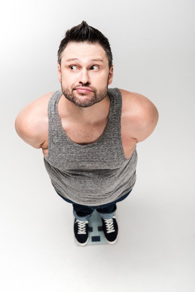высокий угол обзора вдумчивого человека с избыточным весом, стоящего на электронных весах, изолированных на белом
 - Фото, изображение