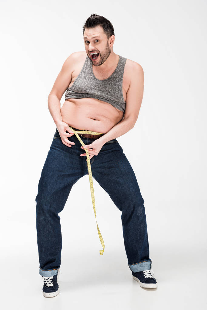 opgewonden overgewicht man kijkend naar camera en meten van de taille met tape op wit - Foto, afbeelding