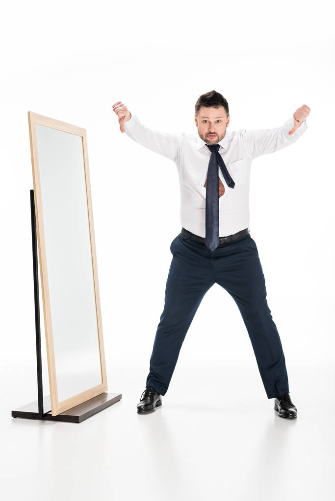homem com sobrepeso no desgaste formal mostrando polegares para baixo perto do espelho no branco
 - Foto, Imagem