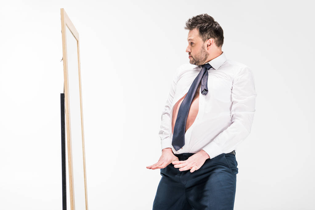 Nadváha muže v těsné formální opotřebování gestikulování a pohledu na zrcadlo izolované na bílém - Fotografie, Obrázek