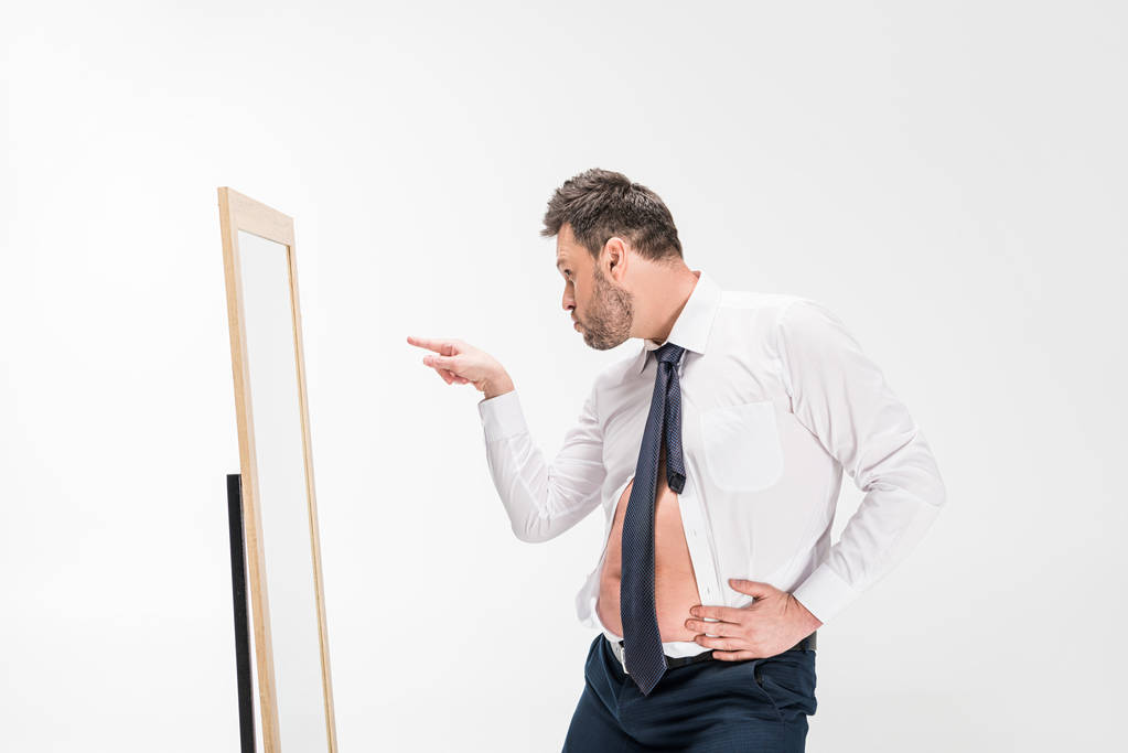 człowiek z nadwagą w formalne zużycie wskazując palcem patrząc na lustro na białym tle - Zdjęcie, obraz