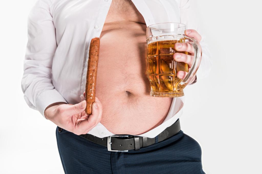 腹を示す太りすぎの男性の部分的なビューと白に隔離されたグリルソーセージとビールのグラスを保持 - 写真・画像