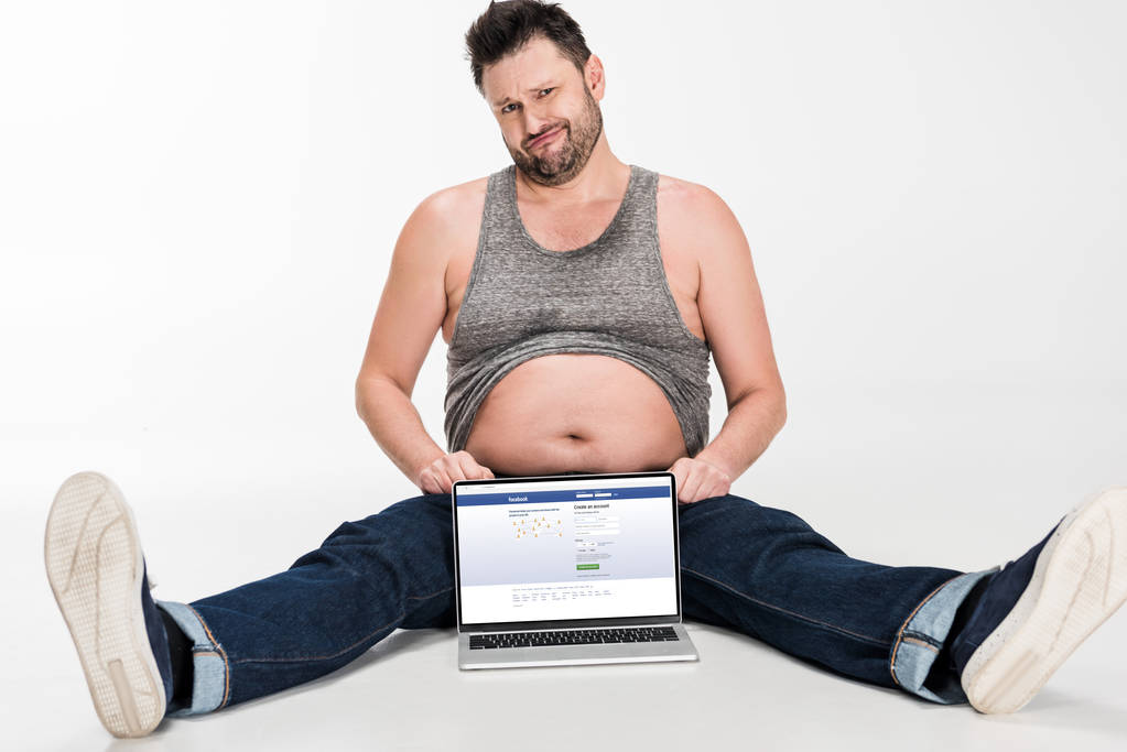skeptical overweight man making facial expression and sitting with laptop with facebook website on screen isolated on white - Photo, Image