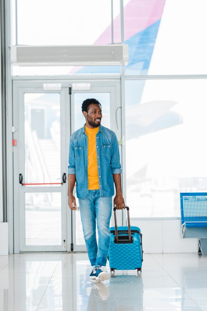 hymyilevä onnellinen afrikkalainen amerikkalainen mies kävely matkalaukku pitkin odotussali lentokentällä
 - Valokuva, kuva