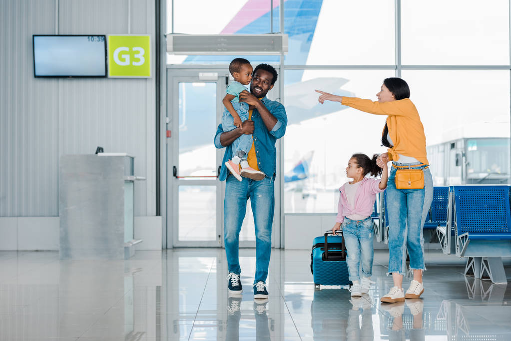 sorrindo família afro-americana andando com bagagem ao longo da sala de espera no aeroporto, enquanto a mãe apontando com o dedo para o filho
 - Foto, Imagem