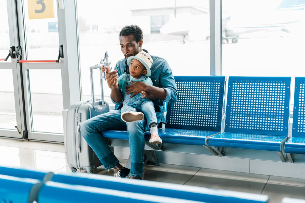 african american father and son sitting in waiting hall in airport and playing with toy plane - Photo, Image