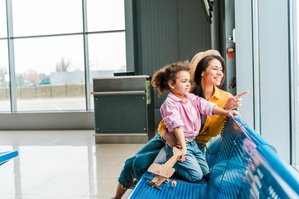 窓に指を指さしながら女性が木製の飛行機モデルで空港に座って幸せなアフリカ系アメリカ人の母と娘 - 写真・画像