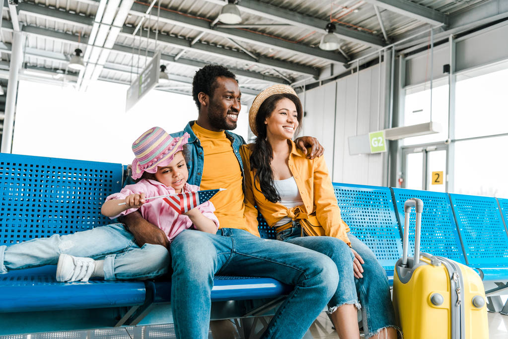 usmívající se africká americká rodina sedící na letišti s kufříkem zatímco dítě drží americkou vlajku - Fotografie, Obrázek