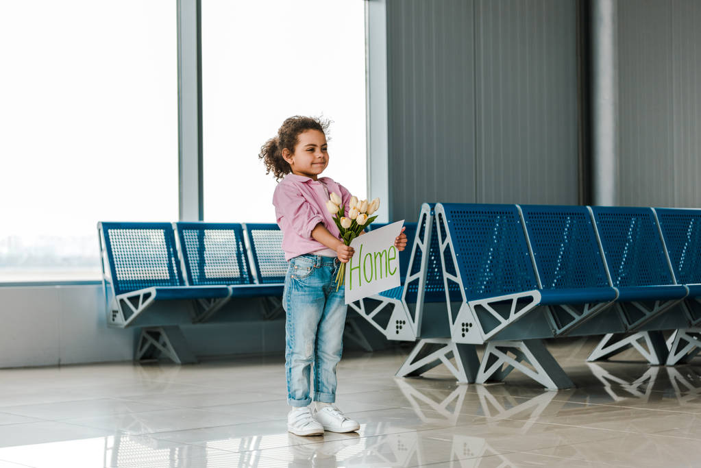 enfant afro-américain joyeux debout dans la salle d'attente à l'aéroport et tenant tulipes et pancarte avec lettrage à la maison
 - Photo, image
