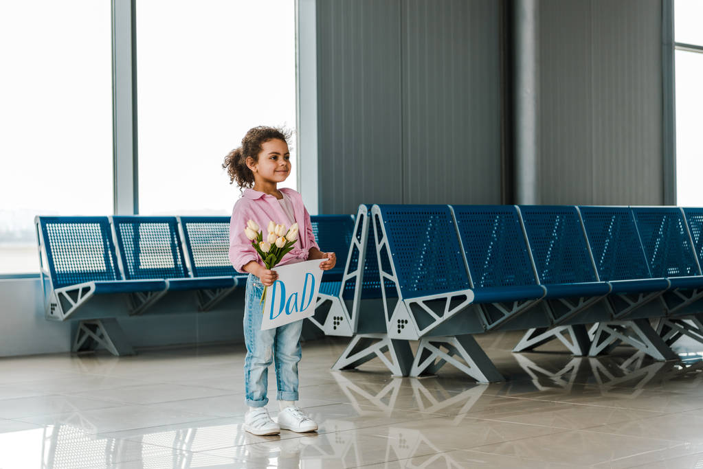 havaalanında bekleyen salonda neşeli Afrikalı Amerikalı çocuk ayakta ve baba yazı ile lale ve plasard tutarak - Fotoğraf, Görsel