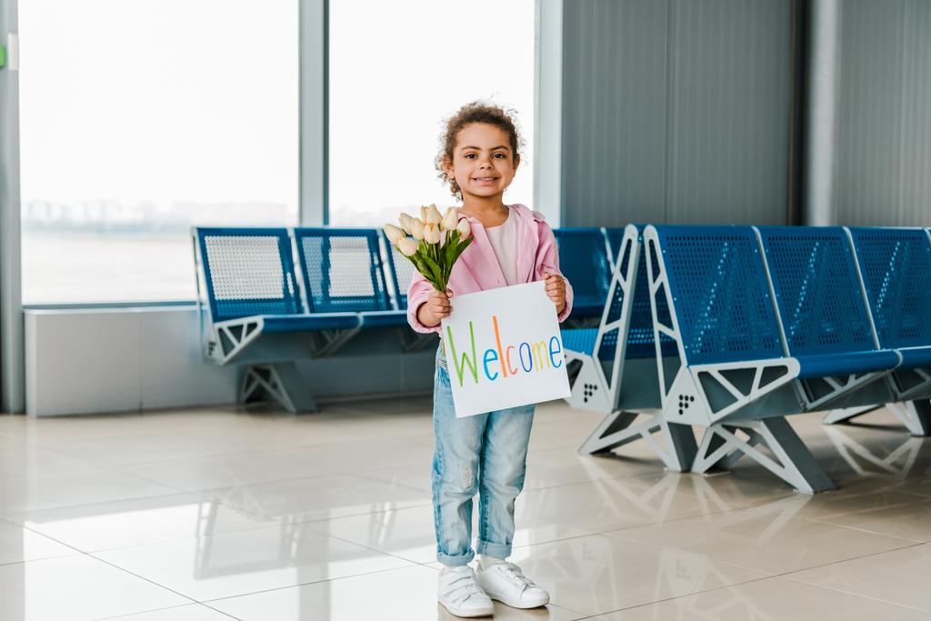 niño afroamericano sonriente parado en la sala de espera en el aeropuerto y sosteniendo tulipanes y pancarta con letras de bienvenida
 - Foto, imagen