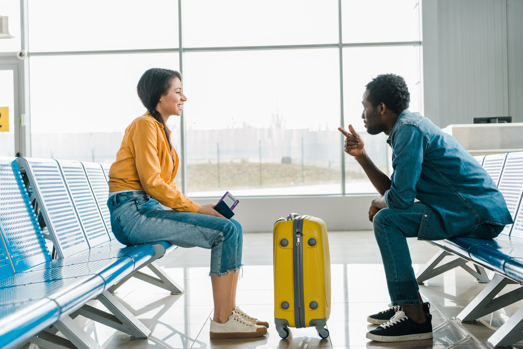 πλευρική όψη του αφρικανικού ζευγαριού που κάθεται στο σαλόνι αναχώρησης με βαλίτσα και αεροπορικά εισιτήρια στο αεροδρόμιο - Φωτογραφία, εικόνα