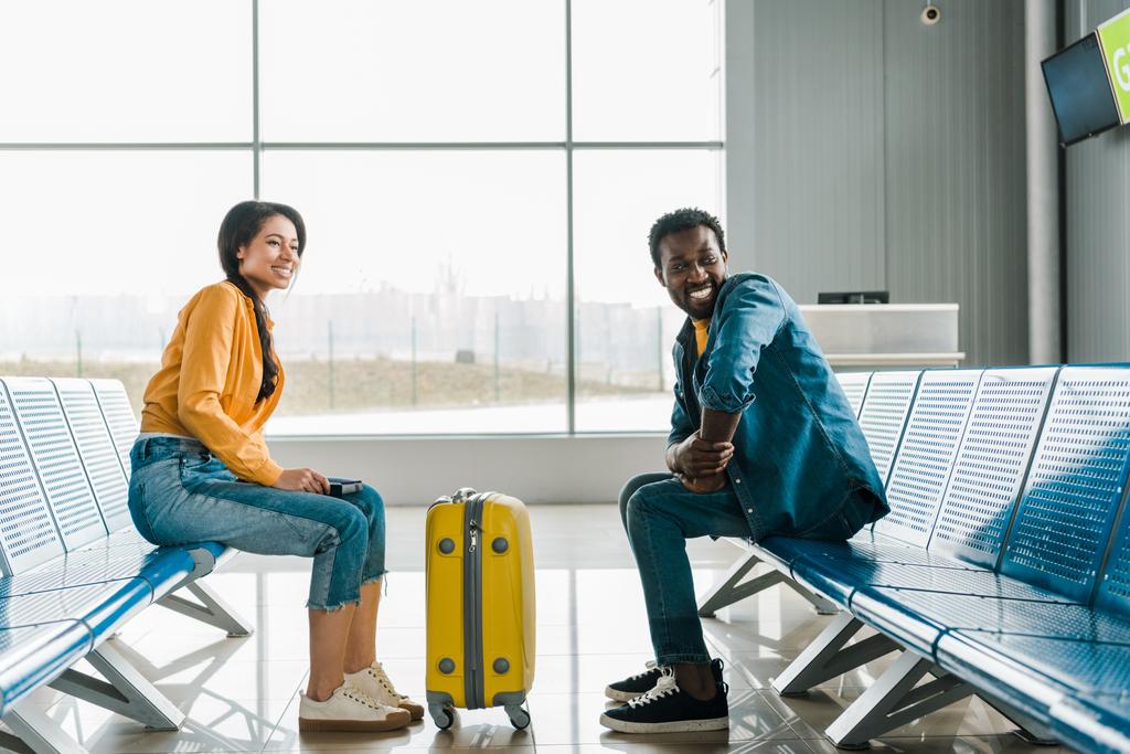 Seitenansicht eines glücklichen afrikanisch-amerikanischen Paares in der Abflughalle mit Koffer am Flughafen - Foto, Bild