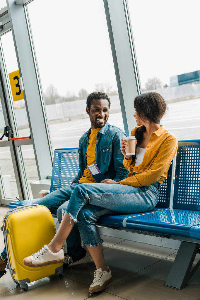 szczęśliwy African American para siedzi w poczekalni z kawą, aby przejść i żółty walizka i patrząc na siebie - Zdjęcie, obraz