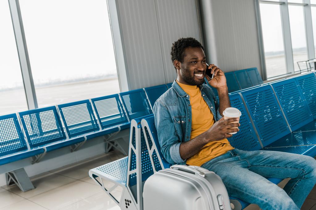 χαμογελαστός Αφρικανός Αμερικανός άνθρωπος κάθεται στο σαλόνι αναχώρησης και μιλάει στο smartphone - Φωτογραφία, εικόνα