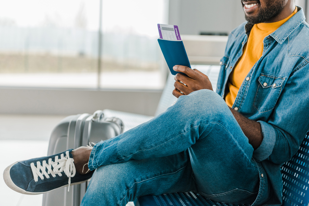 vista ritagliata di sorridente uomo afroamericano seduto in aeroporto con biglietto aereo e passaporto
 - Foto, immagini