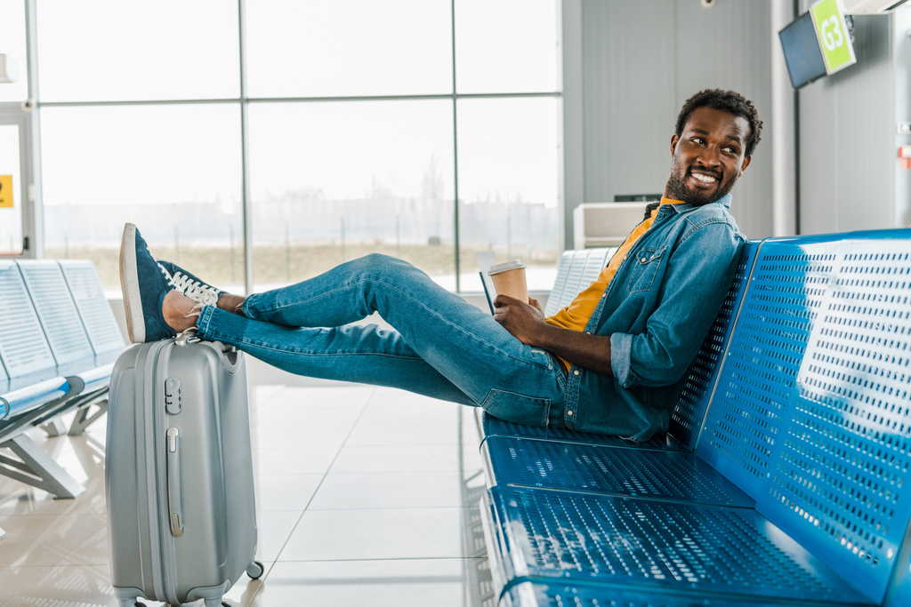 ευτυχισμένος Αφρικανός Αμερικανός άνθρωπος κάθεται στο αεροδρόμιο με καφέ για να πάει και τα πόδια στη βαλίτσα - Φωτογραφία, εικόνα