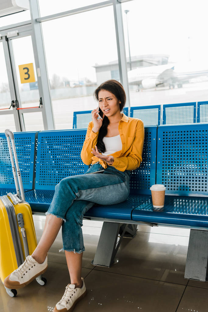 недовольная африканская американка, сидящая в зале вылета с чемоданом, кофе, чтобы пойти и поговорить по смартфону
 - Фото, изображение