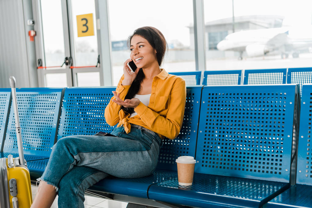 Happy Afro-Amerikaanse vrouw zit in vertrek lounge met koffer, koffie om te gaan en praten op smartphone - Foto, afbeelding