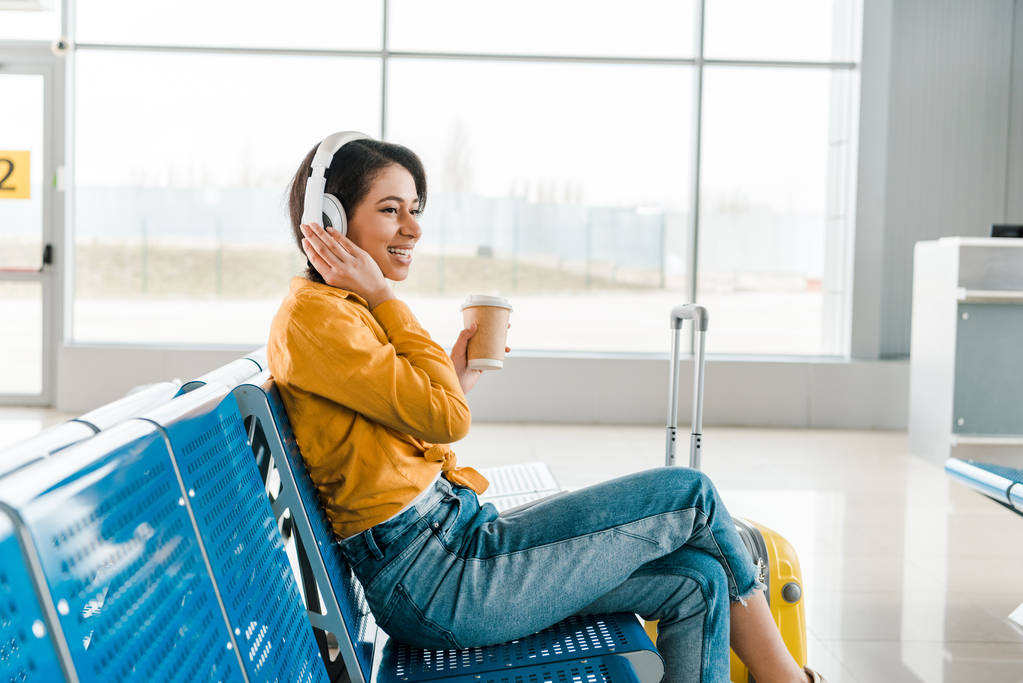 mujer afroamericana feliz sentada en la sala de salida con café para llevar y maleta mientras escucha música en auriculares
  - Foto, Imagen
