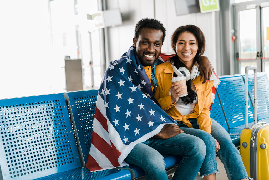 Lächelndes afrikanisch-amerikanisches Paar sitzt mit amerikanischer Flagge in der Abflughalle des Flughafens  - Foto, Bild