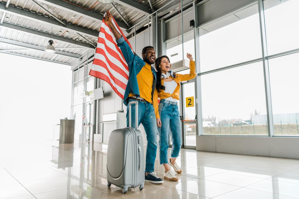 空港の出発ラウンジで歩いているアメリカの国旗とスーツケースを持つハッピー興奮したアフリカ系アメリカ人のカップル  - 写真・画像