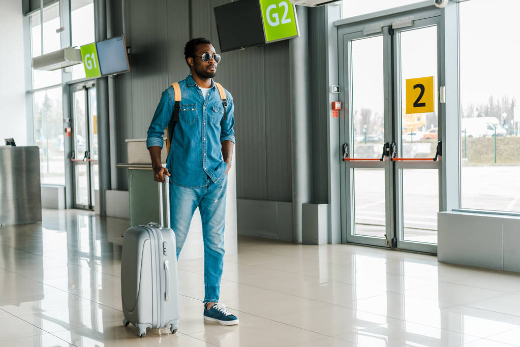 красивий афроамериканський чоловік стоїть з рукою в кишені і валізі в аеропорту
 - Фото, зображення
