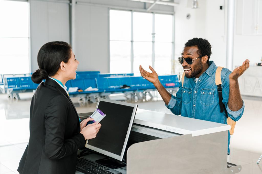 pasajero afroamericano sonriente mostrando gesto de encogimiento mientras el trabajador del aeropuerto sostiene pasaporte y billete de avión
 - Foto, imagen