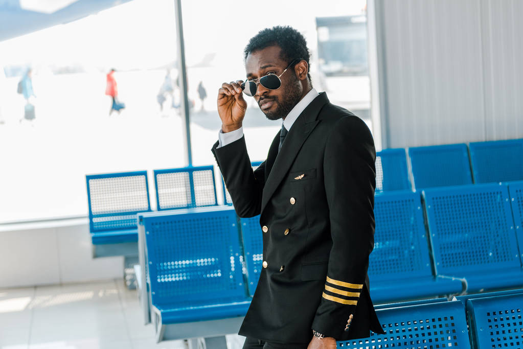 tyylikäs afrikkalainen amerikkalainen pilotti aurinkolaseissa lähellä paikkoja lähtöaulassa lentokentällä
 - Valokuva, kuva