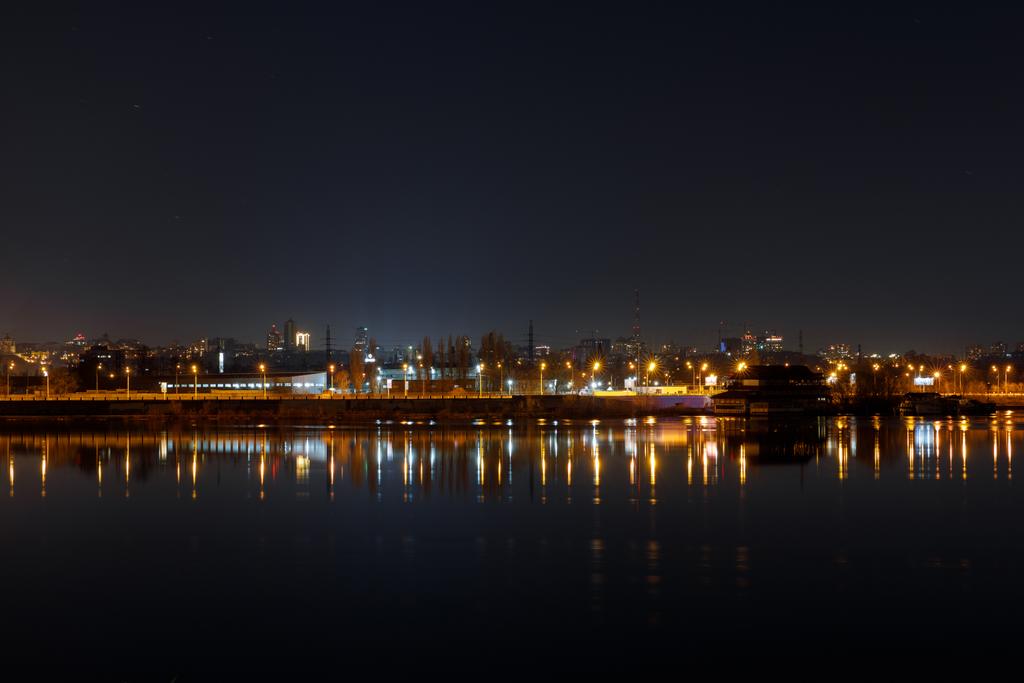 σκούρο αστικό τοπίο με φωτισμένα κτήρια και ήρεμο ποτάμι στο - Φωτογραφία, εικόνα