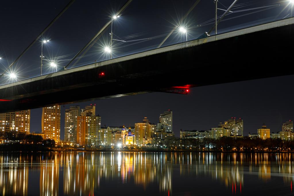 paisaje urbano oscuro con puente, reflexión sobre el río y casas iluminadas por la noche
 - Foto, Imagen