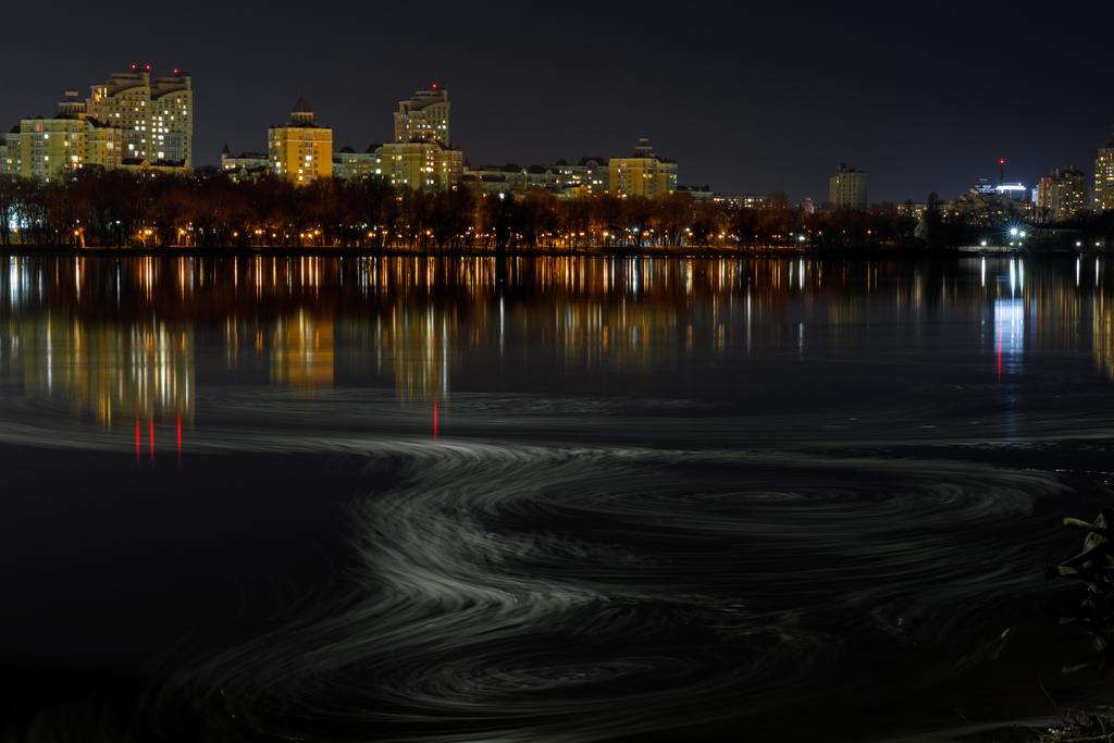 dunkles Stadtbild mit beleuchteten Gebäuden, Wellen am Fluss und Nachthimmel - Foto, Bild