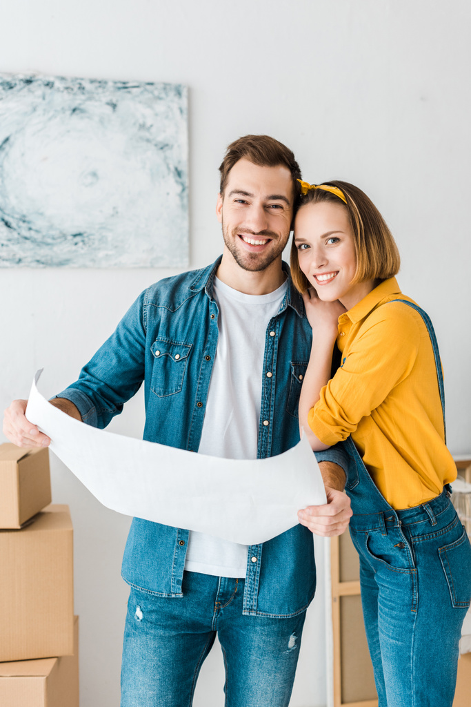 laughing couple in jeans holding blueprint and looking at camera at home - Photo, Image