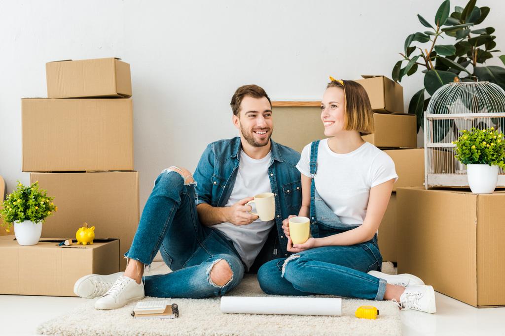 Joyful couple drinking coffee while sitting on floor near cardboard boxes - Photo, Image