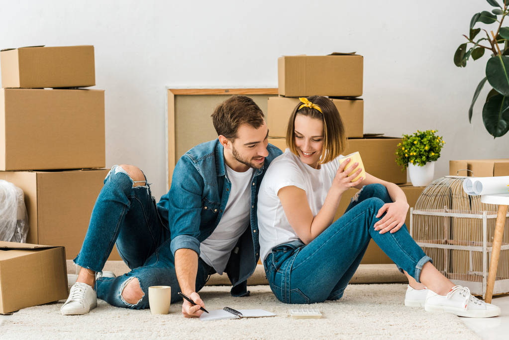smiling couple with notebook sitting on carpet near cardboard boxes and drinking coffee - Photo, Image