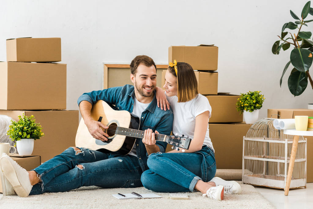 Uśmiechnięty mężczyzna siedzi na dywanie z żoną i grając na gitarze akustycznej - Zdjęcie, obraz