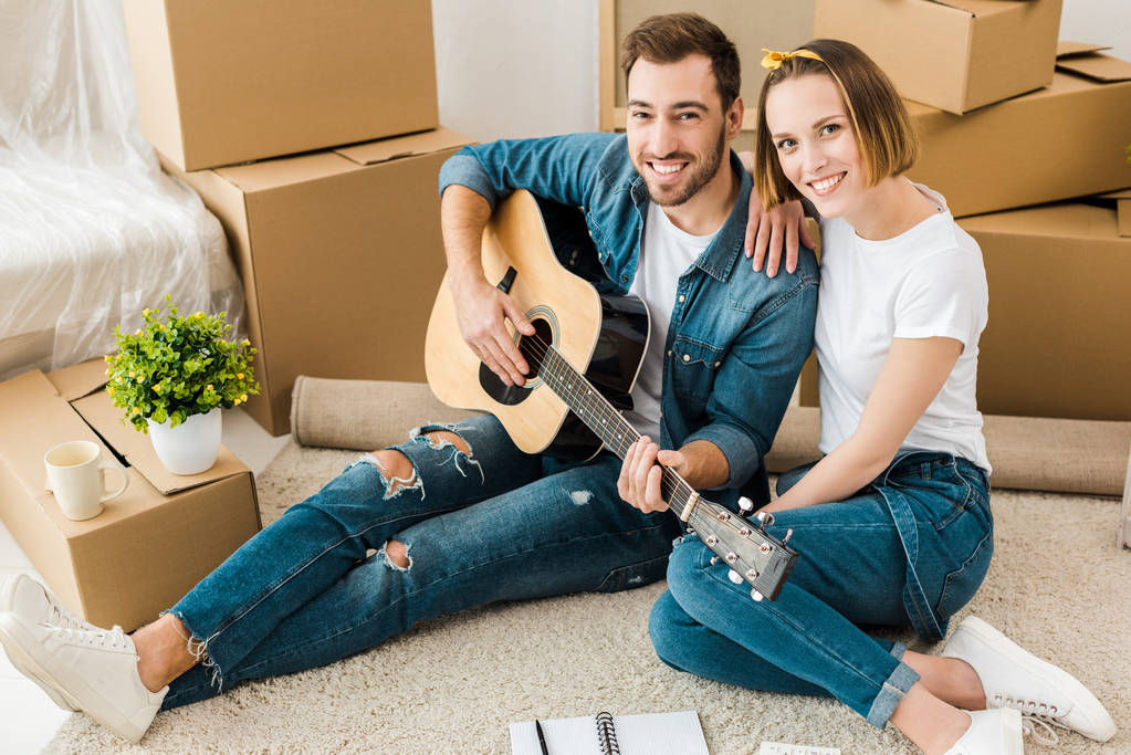 Усміхнений чоловік сидить на килимі з дружиною і грає на акустичній гітарі
 - Фото, зображення