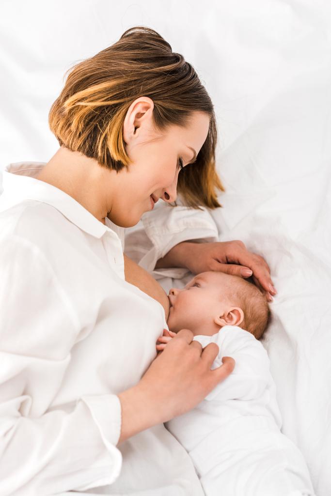 Мать в белой рубашке лежит на кровати и кормит грудью ребенка
 - Фото, изображение