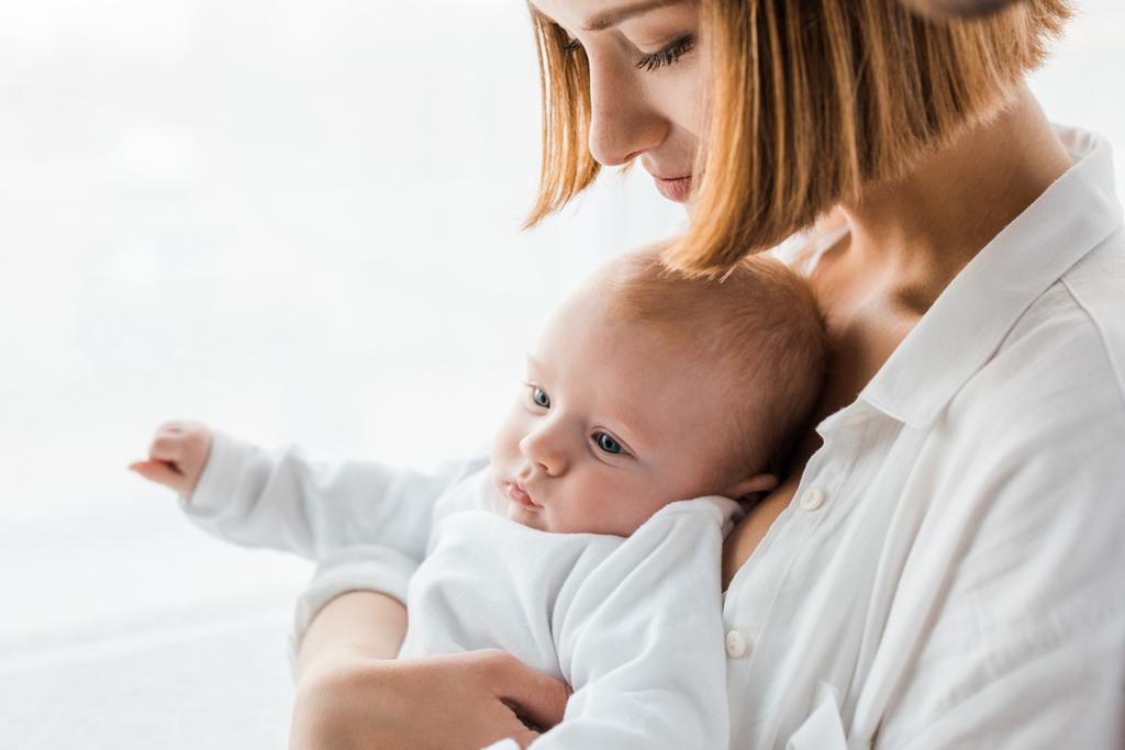 νεαρή μητέρα με λευκό πουκάμισο κρατώντας το μωρό στο σπίτι - Φωτογραφία, εικόνα