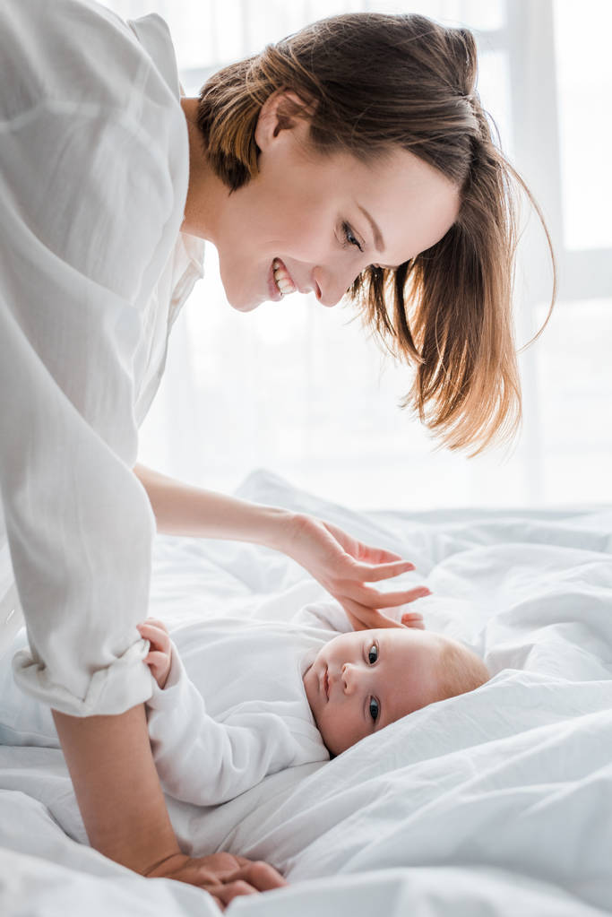 Rindo jovem mãe em camisa branca olhando para o bebê na cama
 - Foto, Imagem